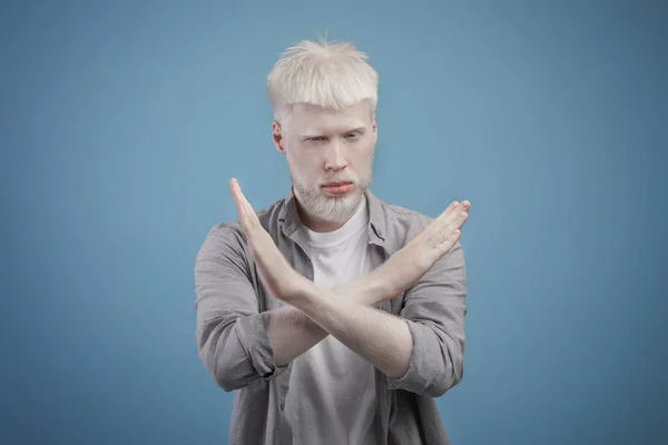 Grave jovem albino cara dizendo não, olhando para a câmera e mostrando as mãos cruzadas sobre o peito, fundo azul — Fotografia de Stock