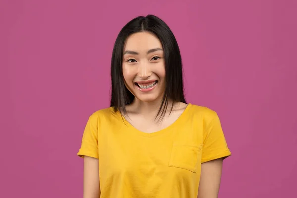 Retrato de bonito ásia mulher no amarelo t-shirt posando sobre violeta estúdio fundo, olhando para câmera e sorrindo — Fotografia de Stock