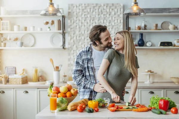 Laki-laki european muda yang puas dengan janggut memeluk perempuan, menyiapkan makanan di meja dengan sayuran organik di dapur — Stok Foto