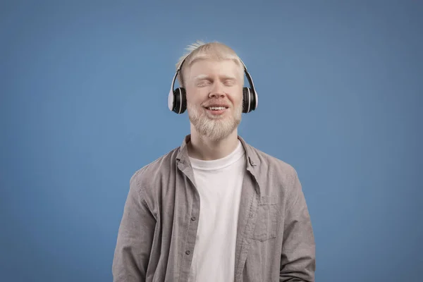 Tevreden rustige albino kerel in hoofdtelefoon genieten van favoriete muziek met gesloten ogen staande over blauwe achtergrond — Stockfoto