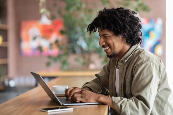 快乐的年轻非洲裔美国卷曲男子在笔记本电脑旁的桌子上打字，桌上有咖啡杯，里面有电话 — 图库照片