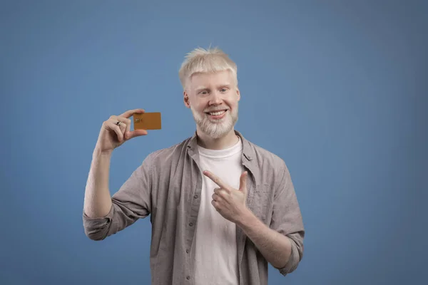 Pria albino bersemangat menunjuk kartu bank di tangannya dan tersenyum di depan kamera, merekomendasikan bank dengan latar belakang biru — Stok Foto