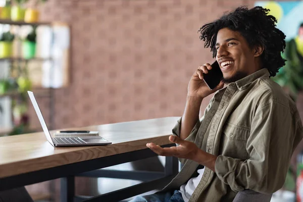 Smiling millennial afrikanisch-amerikanischer Lockenkopf spricht per Smartphone am Tisch sitzend mit Laptop — Stockfoto