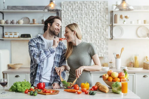 Šťastný mladý evropský manžel s strništěm krmí ženy, dáma připravuje salát k večeři — Stock fotografie