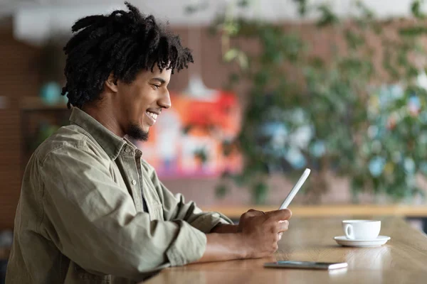 Feliz millennial guapo afroamericano rizado chico blogger escribiendo en la tableta en la mesa con café y teléfono inteligente —  Fotos de Stock