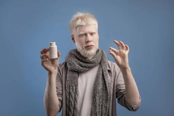 Albino man met pot met medicijnen en pil in de handen, staande op blauwe achtergrond, het nemen van antivirale medicatie — Stockfoto