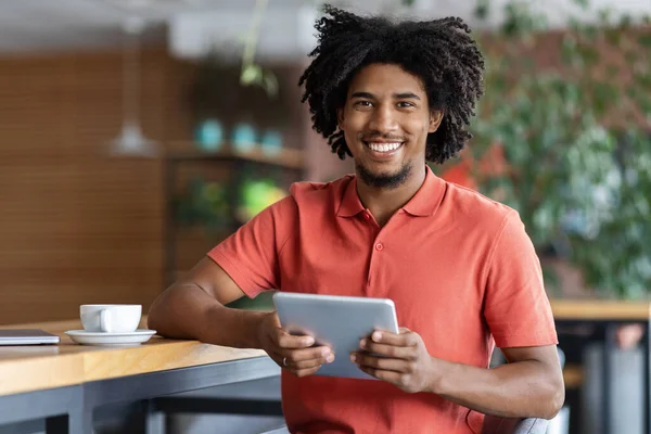 快乐的千年黑人卷曲男子博主在现代咖啡馆内部的平板电脑上与一杯咖啡一起打字 — 图库照片