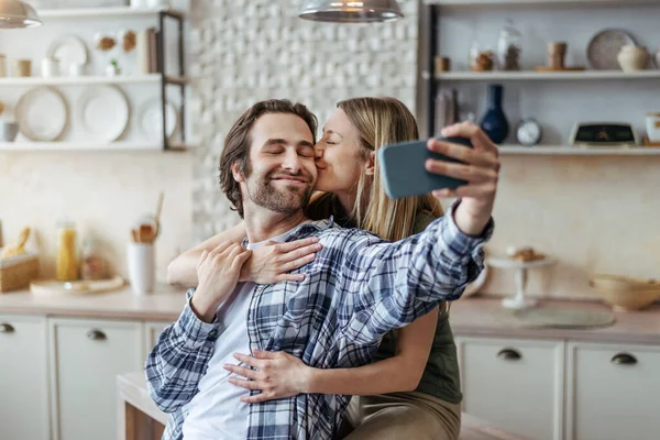 Gülümseyen Avrupalı genç kadın, kocasını sakalla kucaklıyor ve öpüyor, adam akıllı telefondan selfie çekiyor. — Stok fotoğraf