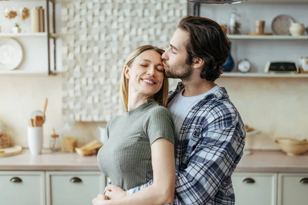 Happy millennial european husband with stubble hugs wife behind, kisses at forehead in kitchen interior — Stock Photo, Image