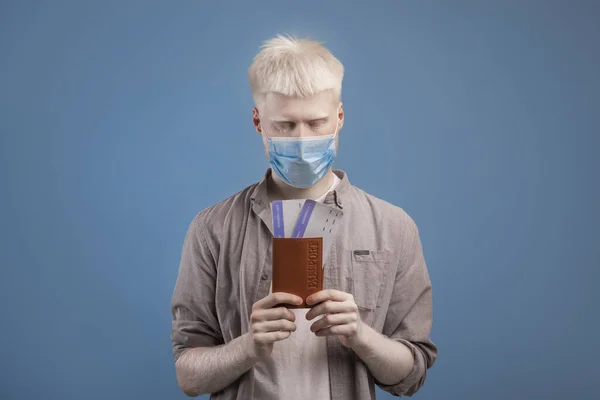 Millennial albino ragazzo indossa maschera facciale, in possesso di passaporto internazionale e biglietti aerei, in posa su sfondo blu — Foto Stock