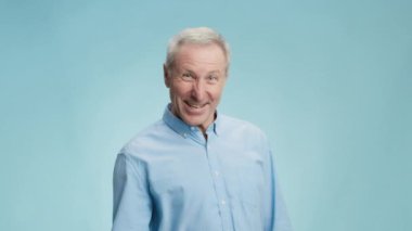 Gömlekli mutlu kıdemli adam yumruğuyla EVET işareti yapıyor, kişisel başarısının tadını çıkarıyor, kameraya gülümsüyor, mavi arka plan