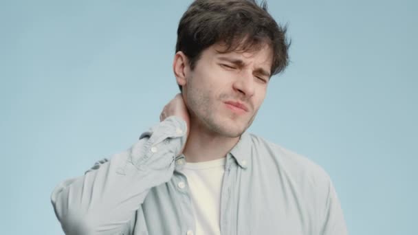Studioporträt eines jungen kranken Mannes mit Nackenschmerzen, massierendem schmerzhaften Körperteil und grimmiger Fratze, blauer Hintergrund — Stockvideo