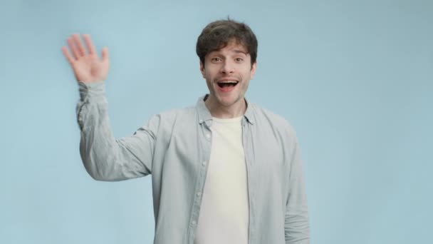 Ahoj. Mladý společenský šťastný muž mává rukou ke kameře, pozdravuje upřímně a široce se usmívající, modré studio pozadí — Stock video