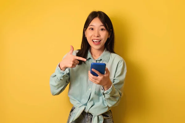Fröhliche junge Asiatin in lässigem Outfit zeigt auf Smartphone-Bildschirm und lernt Online-Verkauf auf gelbem Hintergrund kennen — Stockfoto