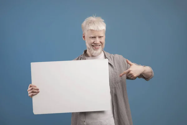 Tempat untuk iklan Anda. Pria albino yang bersemangat memegang papan kosong dan menunjuk ke sana, berpose dengan latar belakang biru — Stok Foto