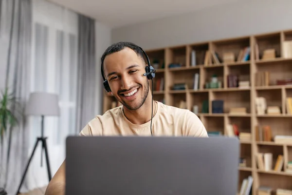 Cheerful middle eastern guy having video chat via laptop wearing wireless headphones, looking at screen and smiling — 스톡 사진