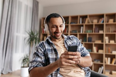 Kablosuz kulaklıklı yakışıklı genç adam evde akıllı telefondan müzik dinliyor.