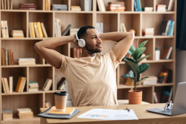 Relaxed arab man listening to music in headphones, resting with hands behind head while working on laptop at home — ストック写真