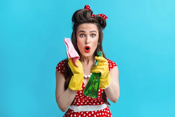 Shocked young pinup woman in retro outfit holding rag and spray detergent, doing domestic work on blue background — стоковое фото