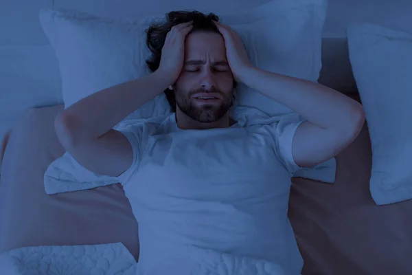 Sleepless guy laying alone in bed, touching his head