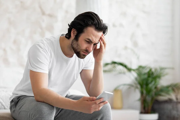 Verärgerter junger Mann mit Smartphone im Bett — Stockfoto