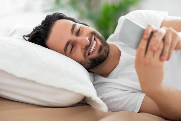 Relaxed guy chatting with friends on smartphone, lying on bed — стоковое фото