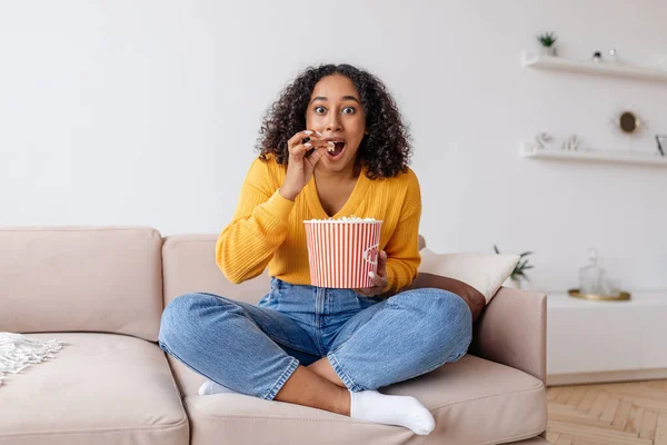 Excited young African American woman sitting on couch, watching TV and eating popcorn at home — Stock Photo, Image