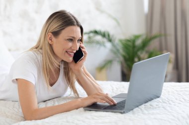Yatakta defter ve akıllı telefon kullanan neşeli bağımsız kadın müteahhit.