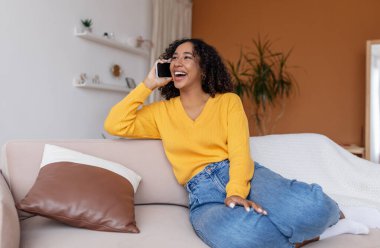 Afro-Amerikan kadın akıllı telefonla konuşuyor, evdeki kanepede oturuyor.
