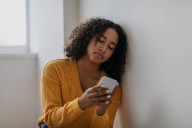 Mutsuz genç siyahi kadın cep telefonundan korkunç haberler okuyor, evde aşırı internet kullanıyor.