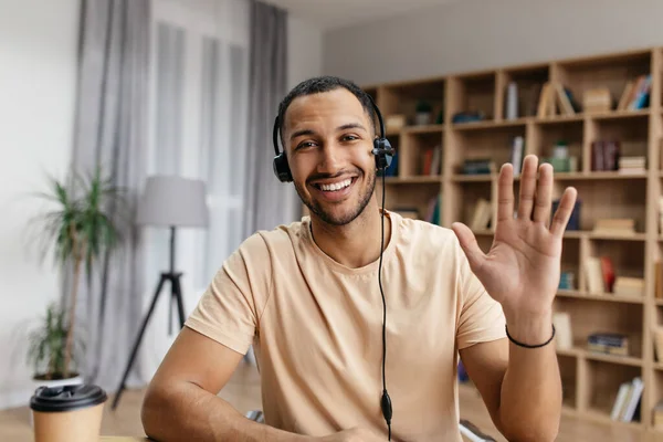 Retrato de profesores en línea. Feliz árabe hombre con auriculares, el gesto de hola a la webcam y sonriendo mientras se sienta en casa —  Fotos de Stock