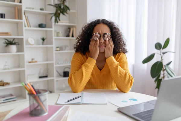 Νεαρή μαύρη γυναίκα με γυαλιά τρίβει τα ερεθισμένα μάτια, κάθεται στο γραφείο με το laptop, μελετώντας online πάρα πολύ στο σπίτι — Φωτογραφία Αρχείου