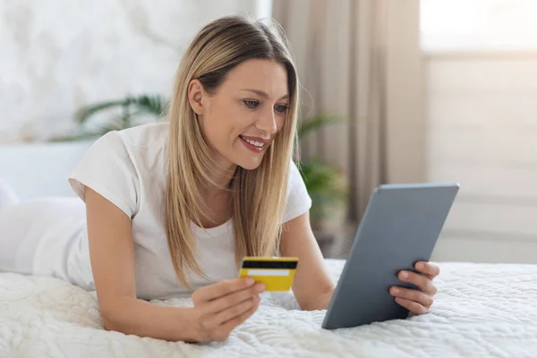 Mujer positiva acostada en la cama con tableta y tarjeta de crédito — Foto de Stock