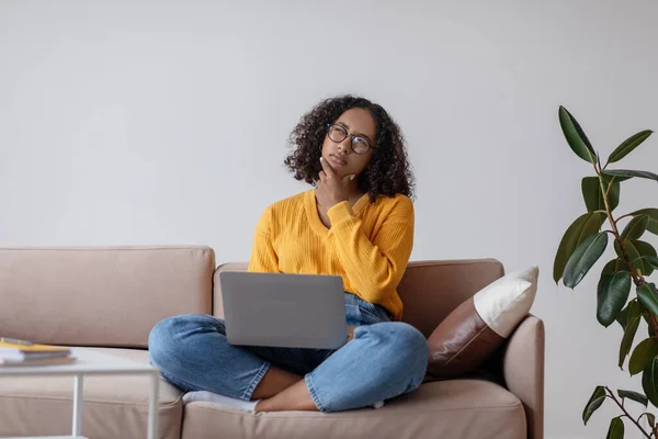 Thoughtful young black female with laptop pc sitting on couch, working online from home office, copy space — Fotografia de Stock