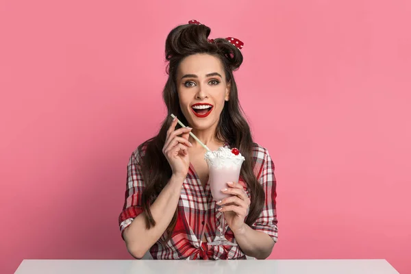 Nadšený mladý pinup žena v retro stylu oblečení pití mléčný koktejl ze skla, sedí u stolu přes modré pozadí — Stock fotografie