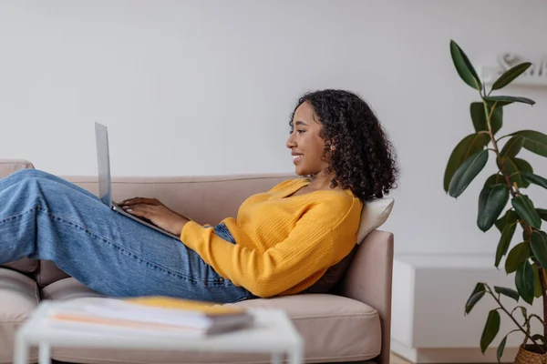Vista lateral da jovem afro-americana trabalhando online via computador portátil na sala de estar — Fotografia de Stock
