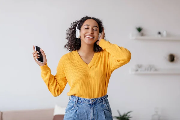 Lovely black woman listening to music in wireless headphones, using smartphone app, enjoying favorite songs at home — Foto Stock