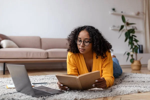 Focalizzato giovane donna nera libro di lettura, sdraiato sul pavimento vicino al computer portatile, studiare online da casa — Foto Stock
