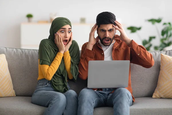 Pareja musulmana conmocionada usando el ordenador portátil sentado en el sofá en casa —  Fotos de Stock