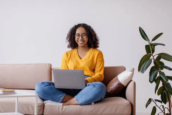 Feliz mulher negra milenar em óculos usando computador portátil, sentado no sofá em casa, espaço de cópia — Fotografia de Stock