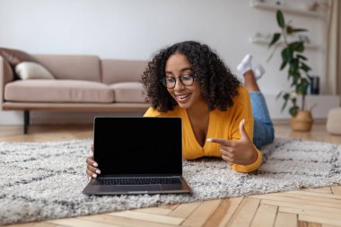 Mutlu genç siyahi kadın evde yerde yatıyor, ekranda web sitesi için dizüstü bilgisayarı gösteriyor.