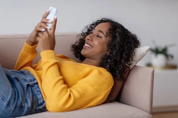 Cheerful young black female communicating online on smartphone, lying on couch in living room — Foto Stock