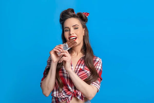 Sexy jovem pinup mulher no retro estilo roupa comer chocolate bar no azul estúdio fundo — Fotografia de Stock