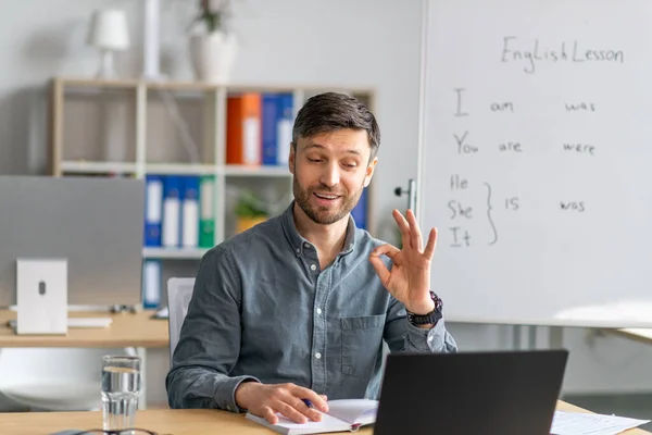 Cheerful male teacher giving online English lesson on laptop computer, showing OK gesture at office interior — Foto de Stock