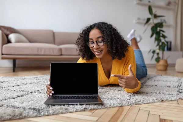 Happy young black woman lying on floor at home, pointing at laptop pc with mockup for website on screen — Foto Stock