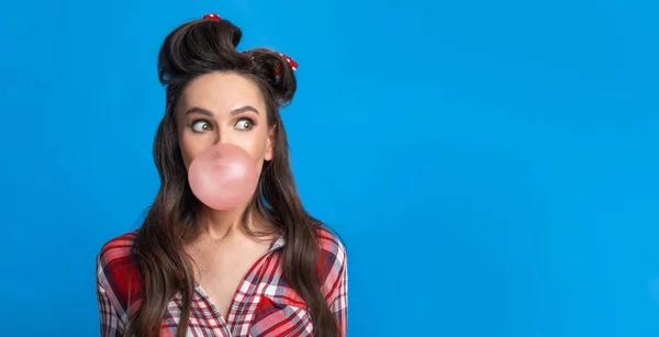 Emocionální tisíciletá pinup žena v retro stylu outfit foukání bublina z žvýkačky přes modré studio pozadí — Stock fotografie
