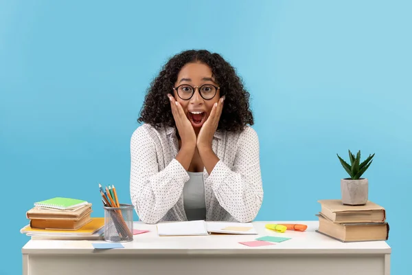 Podekscytowana młoda czarna kobieta w okularach siedząca przy biurku z materiałami do nauki, osiągająca sukces w egzaminach na niebieskim tle — Zdjęcie stockowe