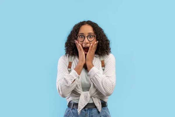 Scared young black female student in glasses wearing backpack, screaming in terror on blue studio background — Stock Photo, Image