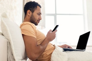 Orta Doğulu Serbest Çalışan Adam Yatak odasında Akıllı Telefon ve Laptop Kullanıyor