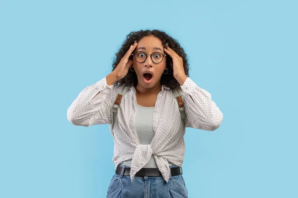 Portrait of shocked young black female student in eyeglasses grabbing her head in terror on blue studio background — Stockfoto